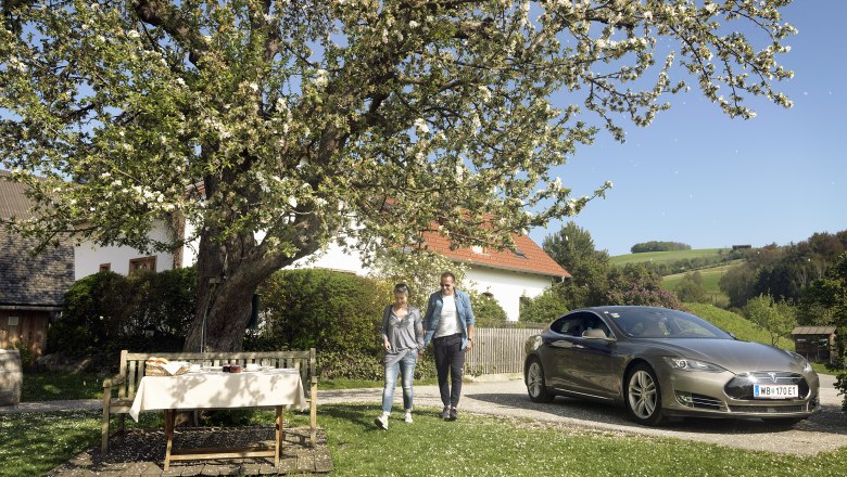 Pár se prochází vedle zaparkovaného elektromobilu pod rozkvetlým stromem ve venkovské zahradě.