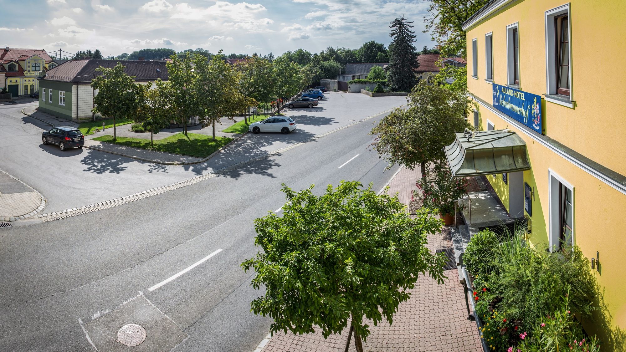Scéna ulice se žlutou budovou a parkovištěm, stromy a auty.