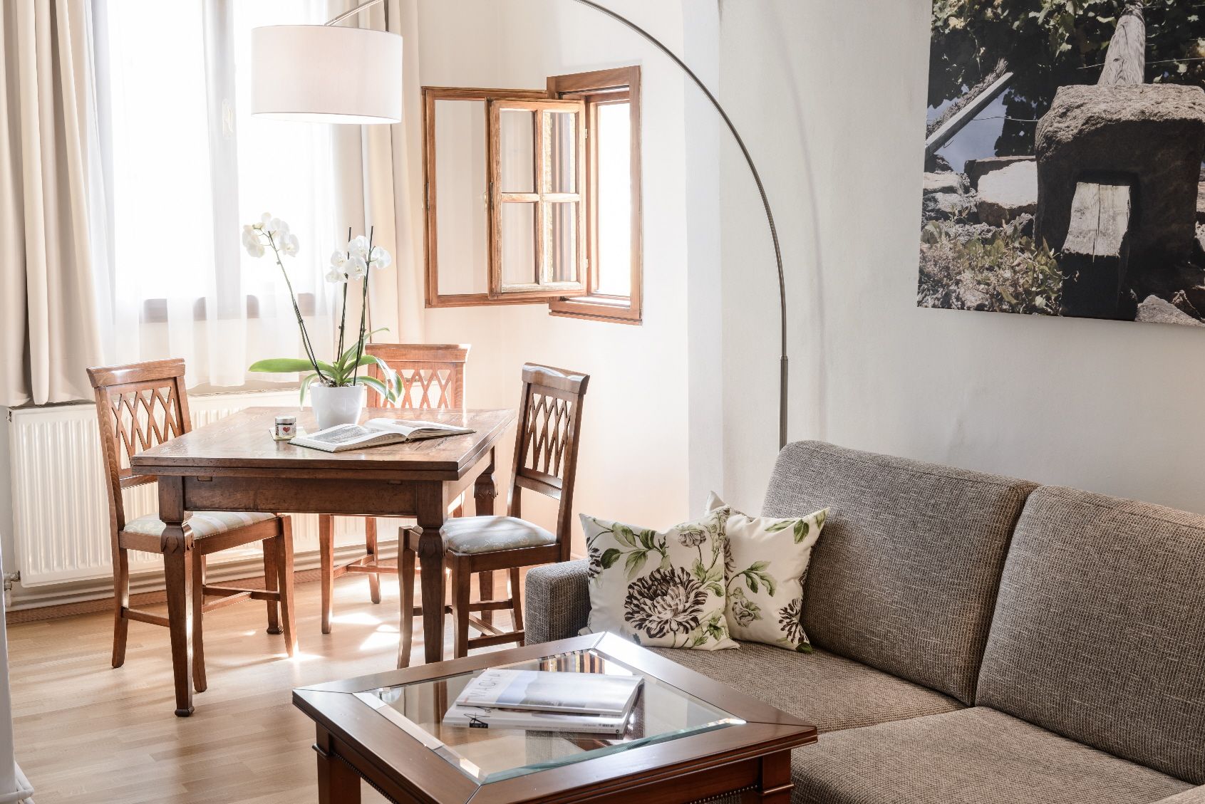 Útulný obývací pokoj s šedou pohovkou, dřevěným stolem a židlemi, světlým oknem a rostlinami.