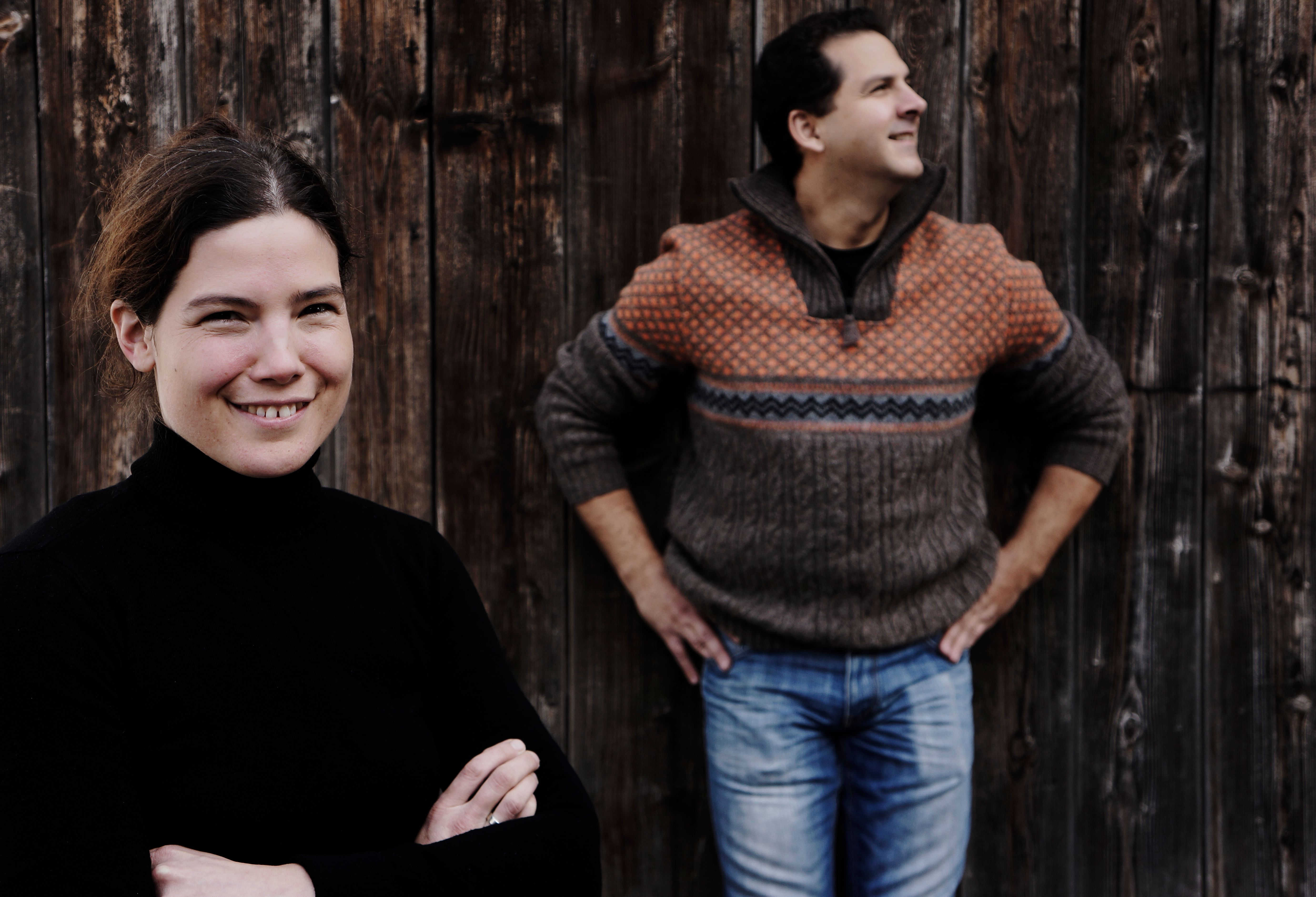 Dva lidé před dřevěnou stěnou, žena v popředí se usmívá, muž v pozadí se dívá stranou.