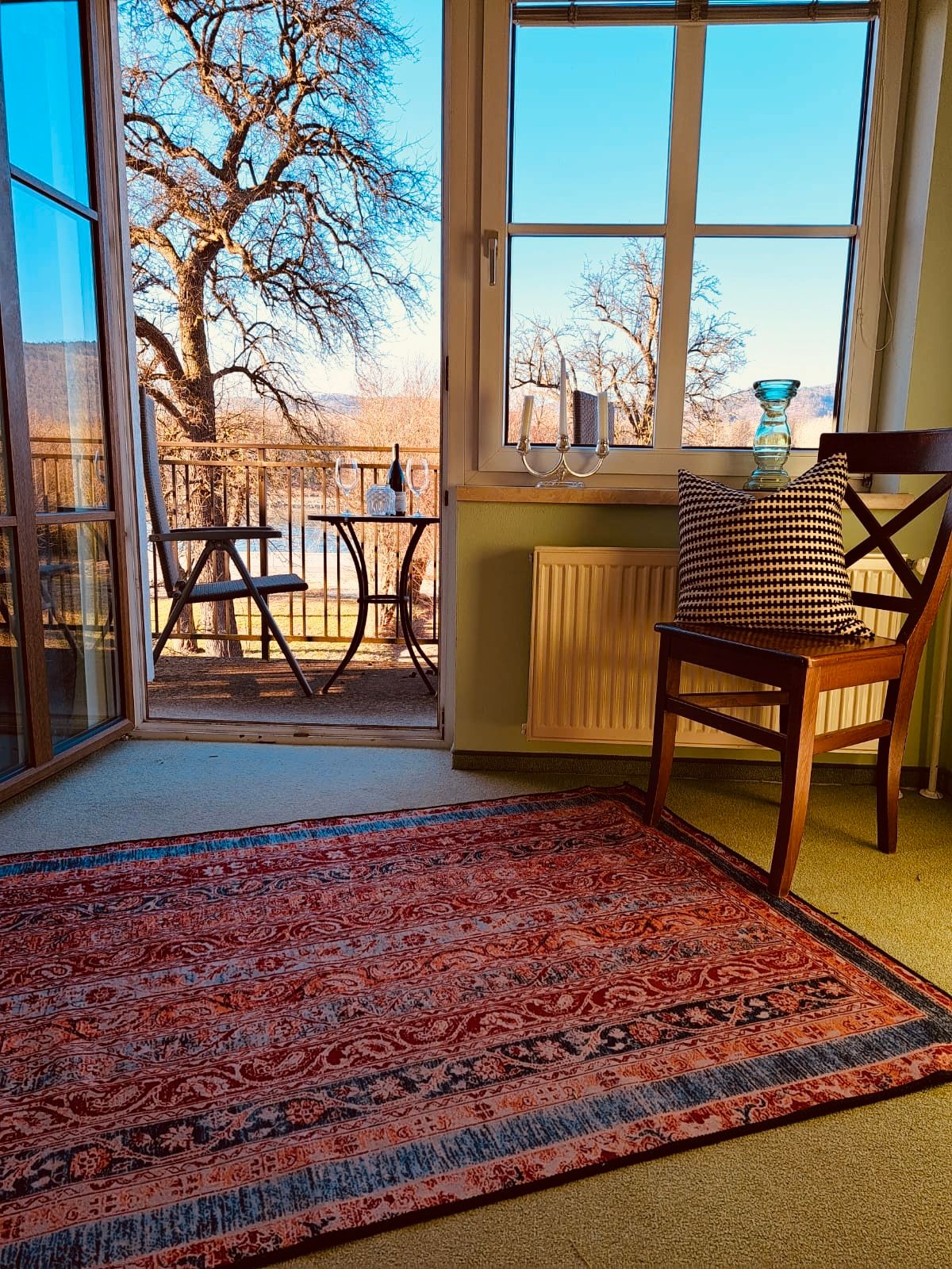 Pokoj s kobercem, židlí a výhledem na balkon se stolem a židlemi, v pozadí stromy.