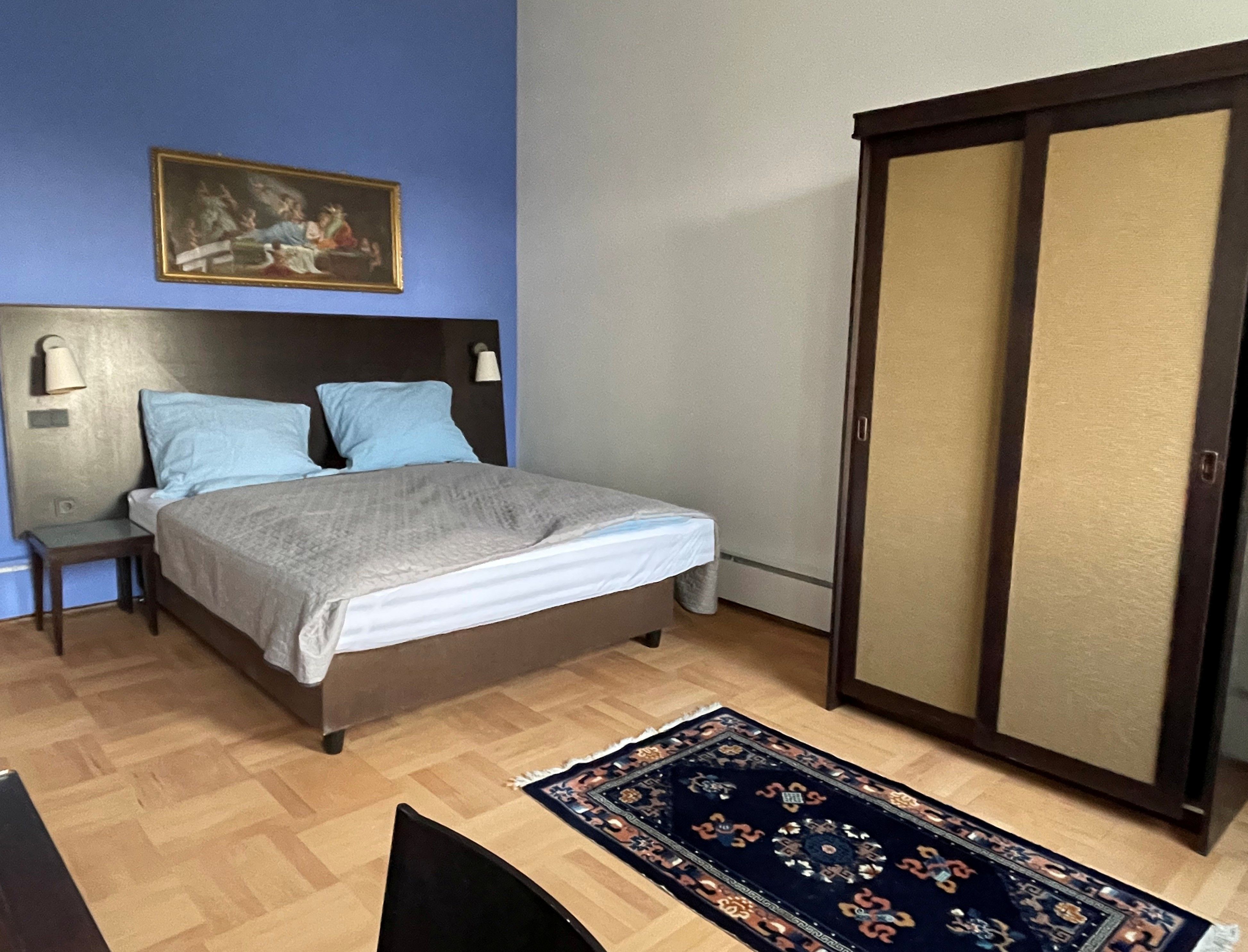 Ložnice s modrou stěnou, manželskou postelí, skříní a kobercem na dřevěné podlaze.