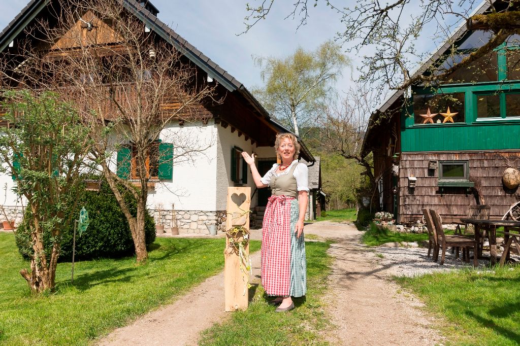 Žena v tradičním oděvu stojí před dvěma venkovskými staveními na zeleném nádvoří.
