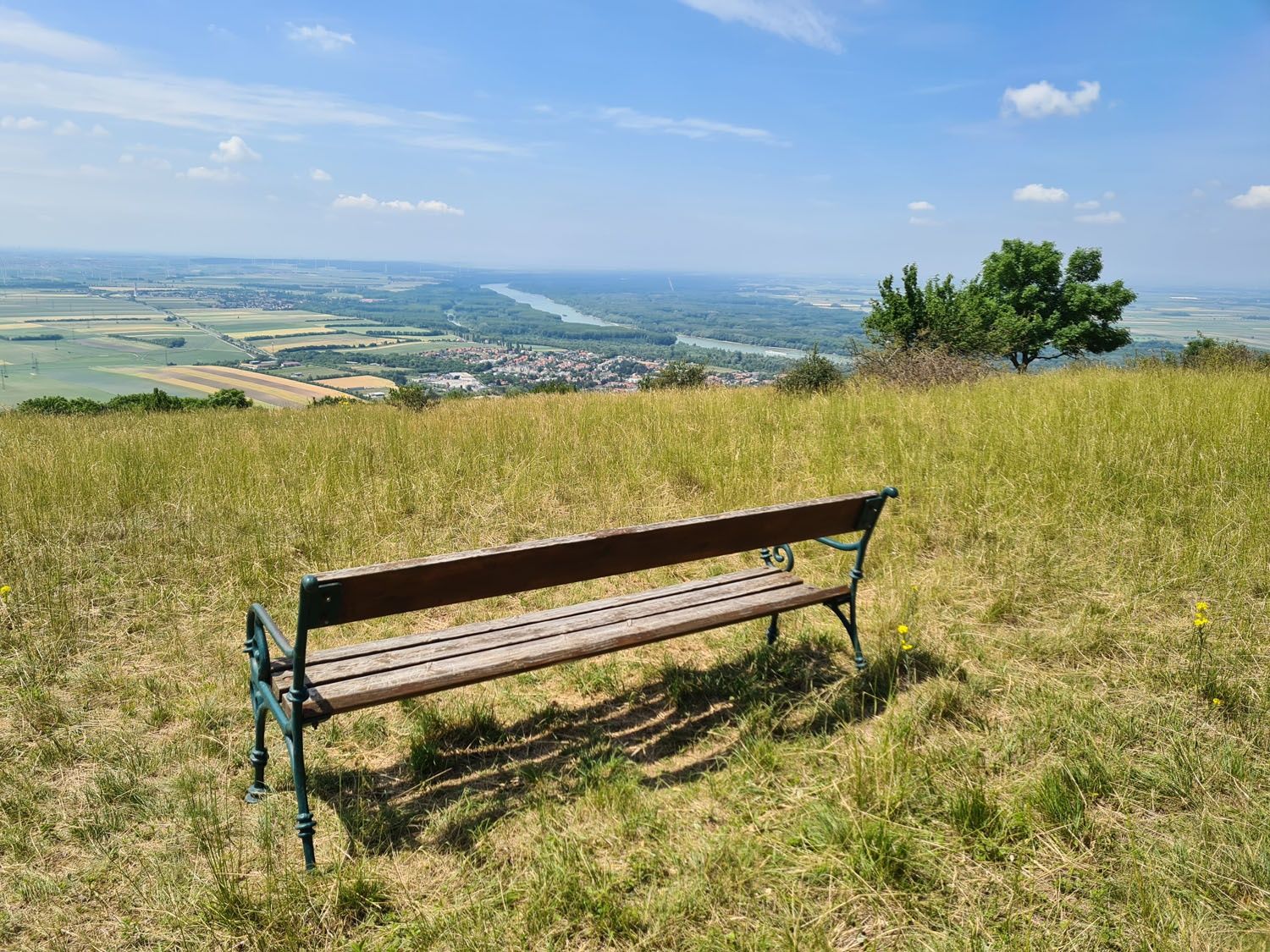 Dřevěná lavička na louce s výhledem na rozlehlou krajinu a řeku v dálce.