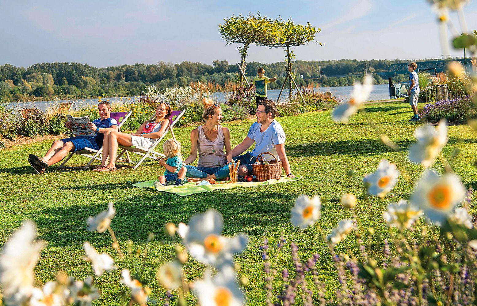 Rodinný piknik v přírodě s květinami v popředí a řekou v pozadí.