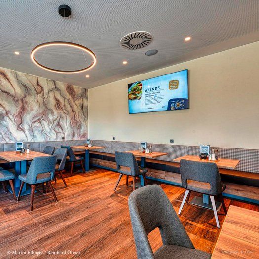 Moderní restaurace s dřevěnou podlahou, šedými židlemi a velkou obrazovkou na stěně.
