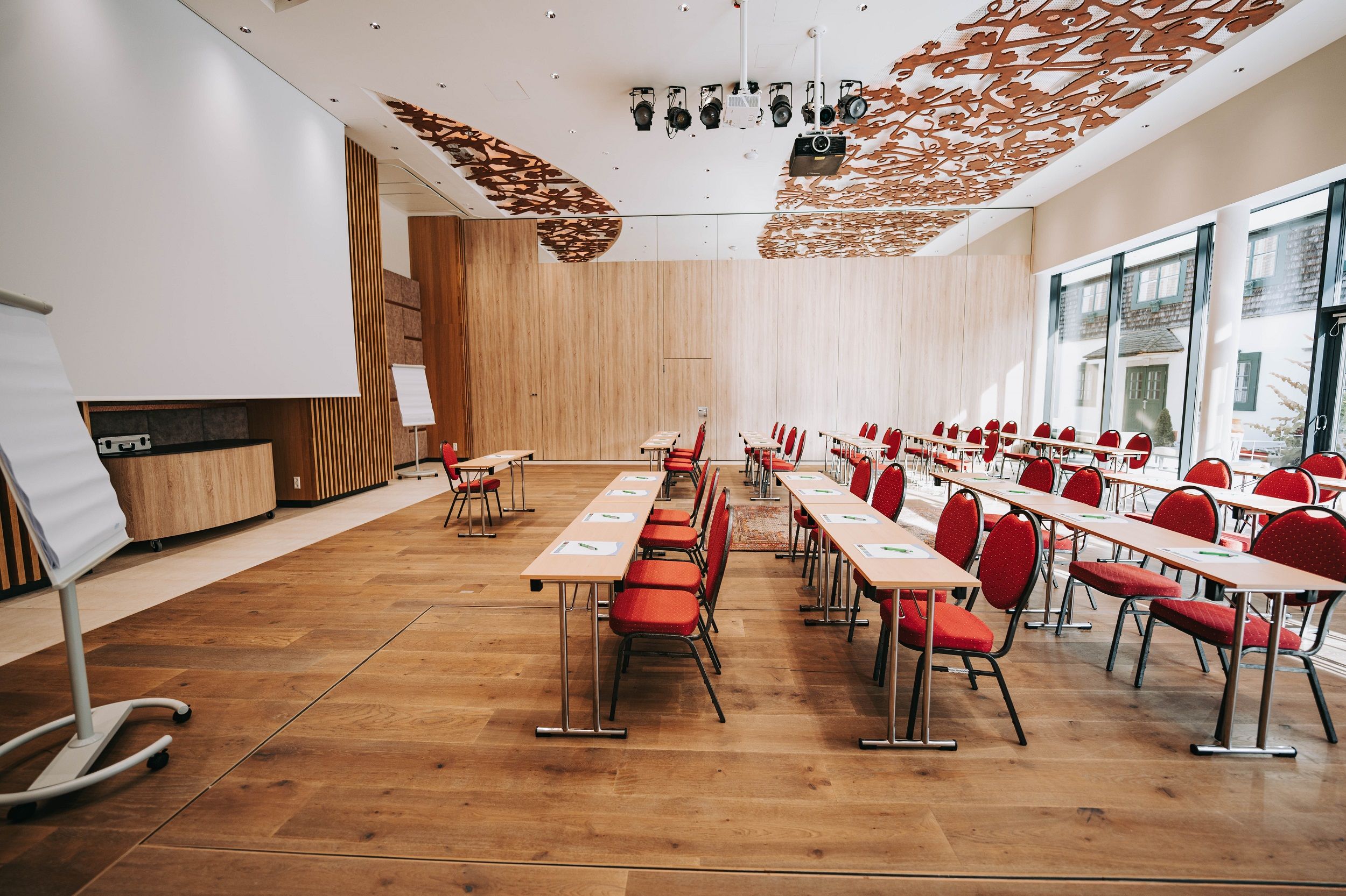 Pohled do interiéru moderní konferenční místnosti s červenými židlemi a dřevěnou podlahou.