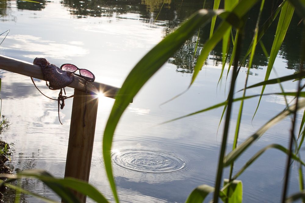 Dřevěné zábradlí na břehu jezera se slunečními brýlemi a bikinami. Ve vodě jsou vidět malé vlnky obklopené rákosím.