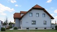 Pohled zvenčí na šedý dům s červenou střechou a několika okny.