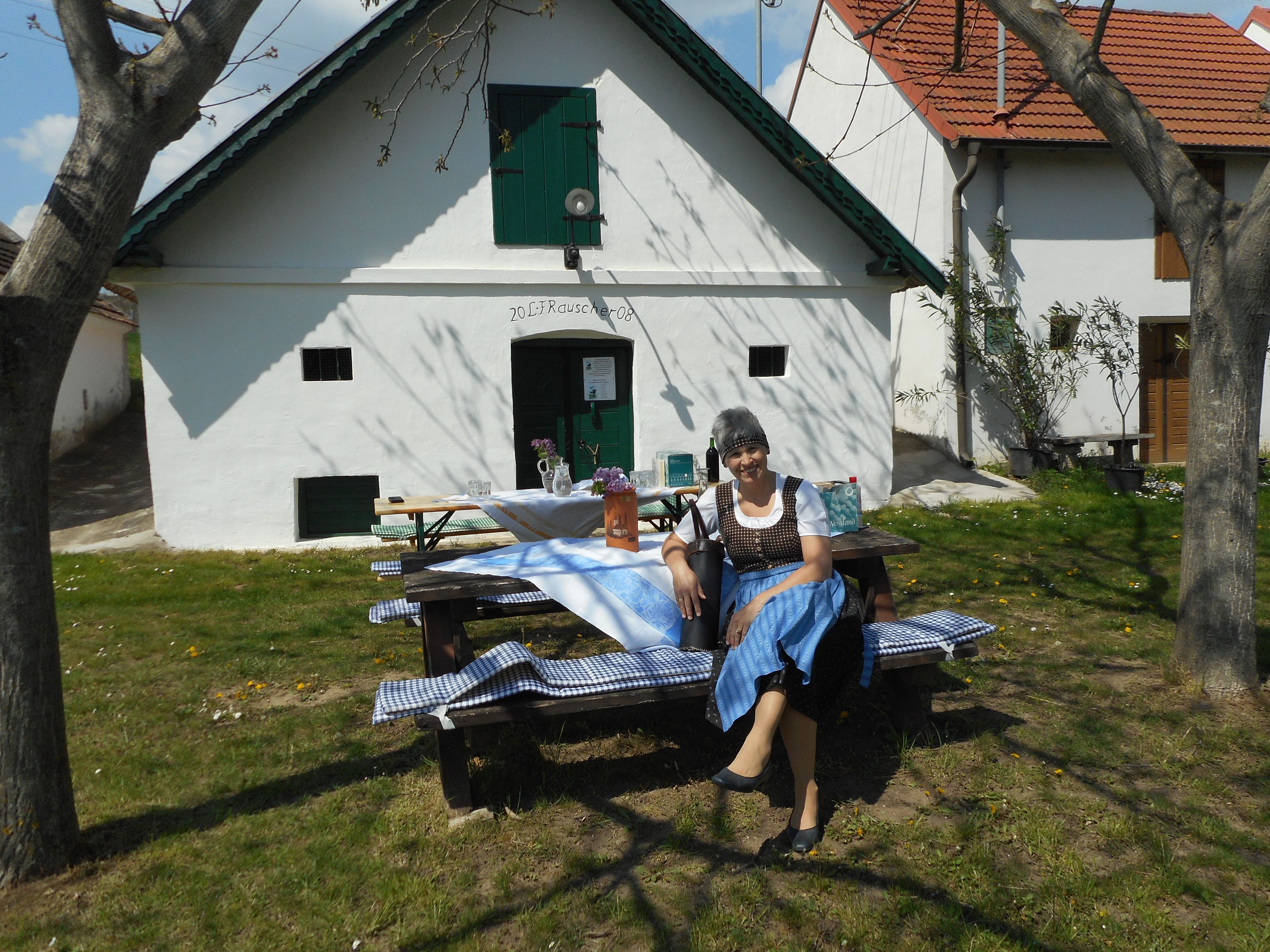 Žena v tradičním oděvu sedí na lavičce před bílou budovou se zelenými okenicemi.