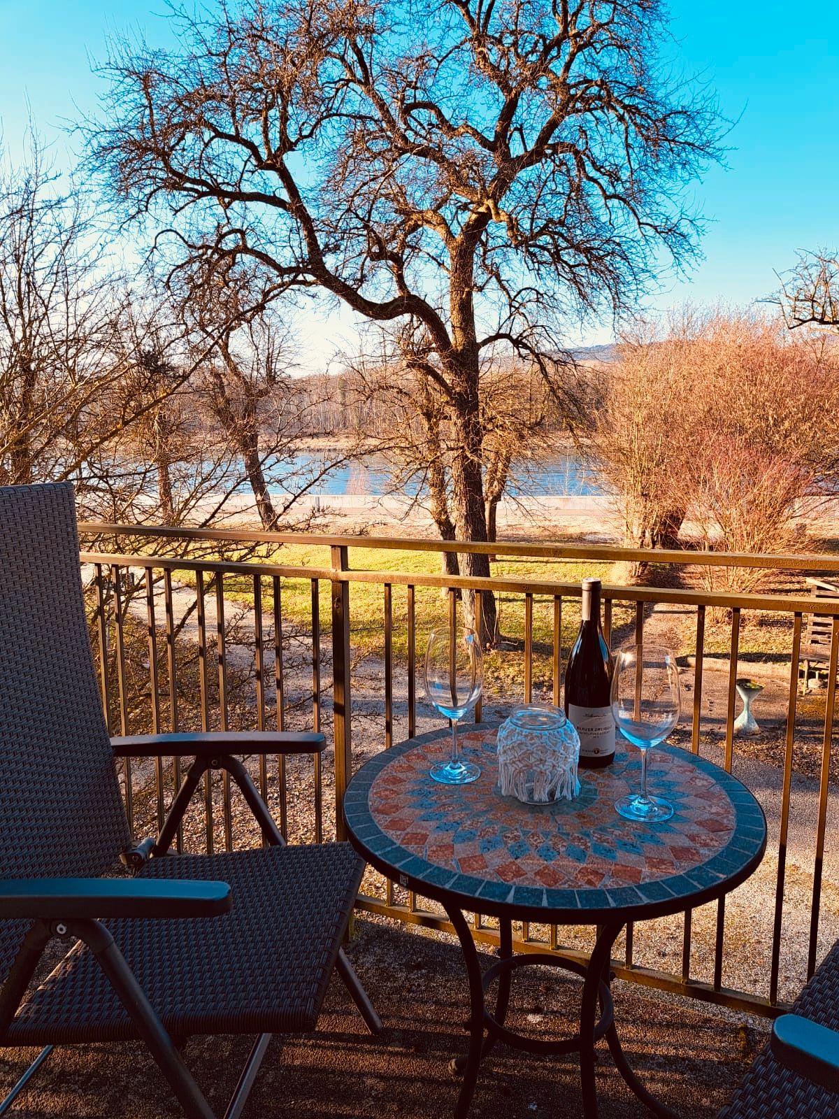Balkon se stolem, dvěma sklenicemi na víno a lahví, výhled na strom a řeku.