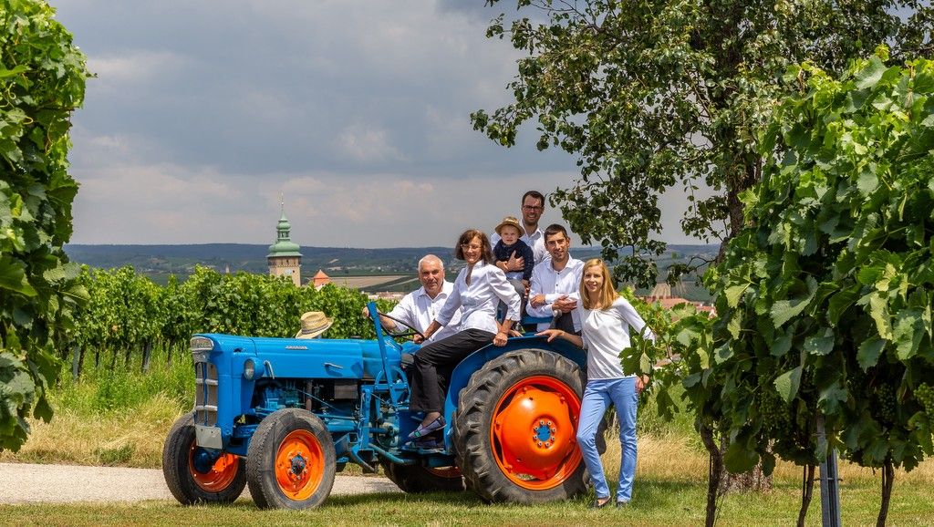 Skupina lidí na modrém traktoru na vinici.