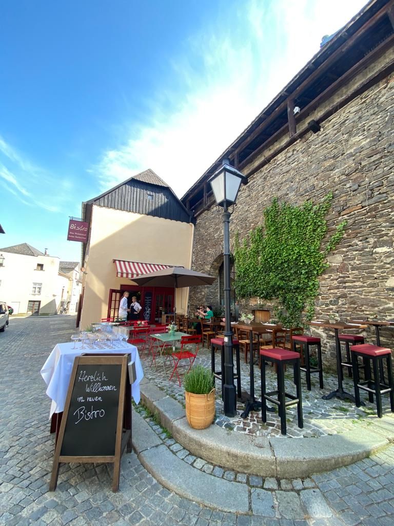Útulné bistro s červenými židlemi a stoly venku, obklopené starými kamennými zdmi a rostlinami.