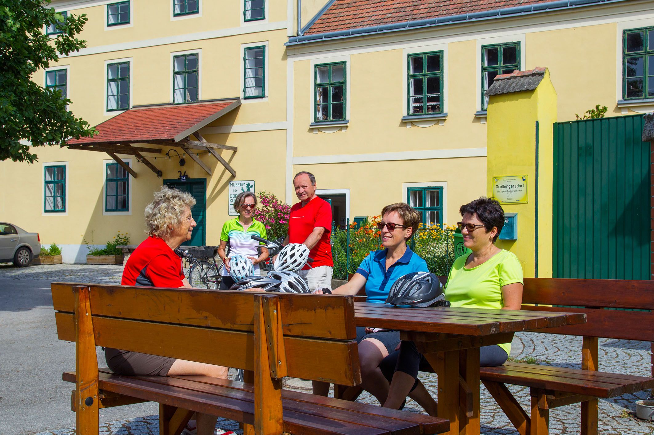 Skupina cyklistů sedících u dřevěného stolu před žlutou budovou.