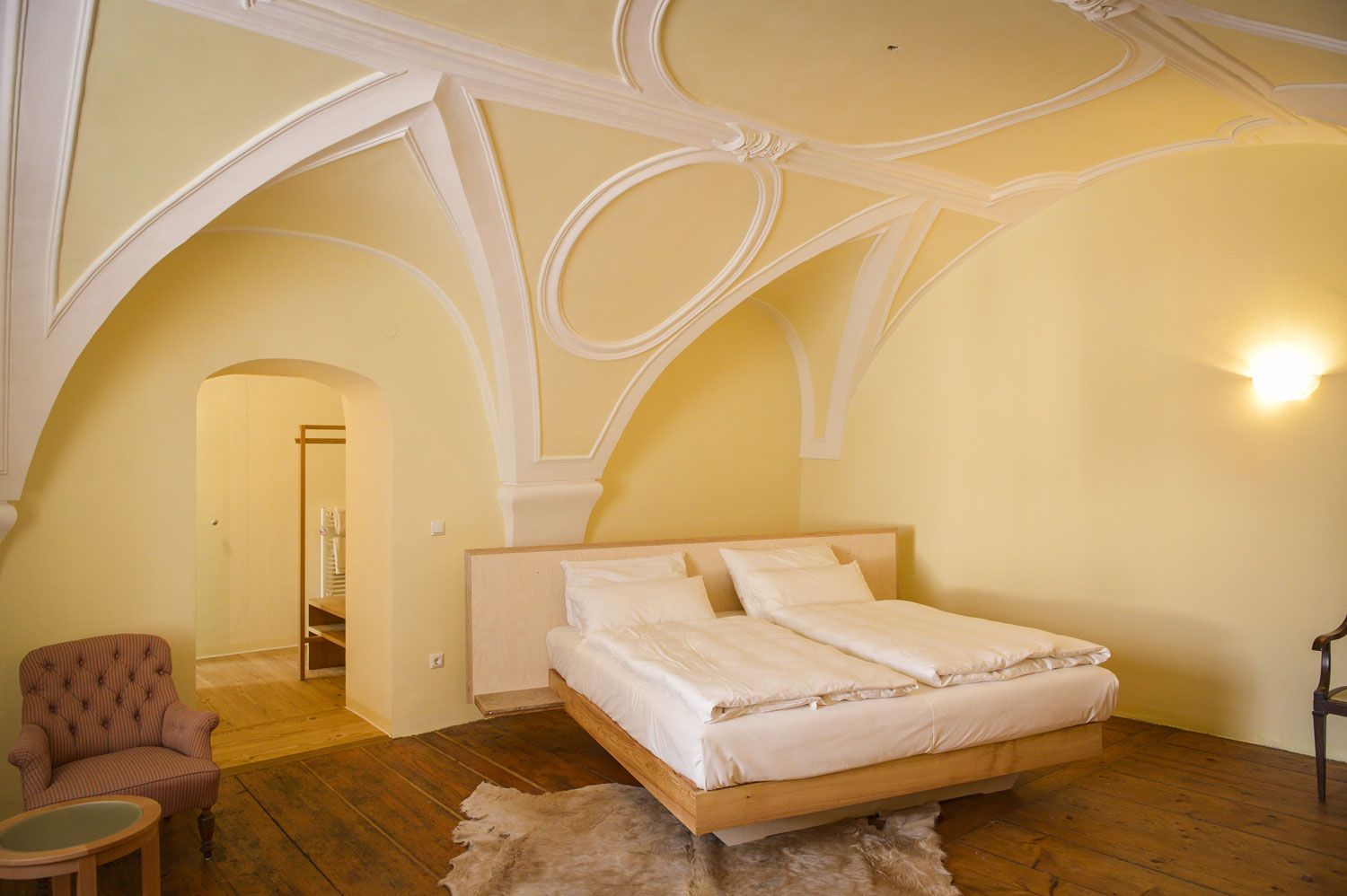 Elegantní ložnice s klenutým stropem, manželskou postelí, křeslem a dřevěnou podlahou.