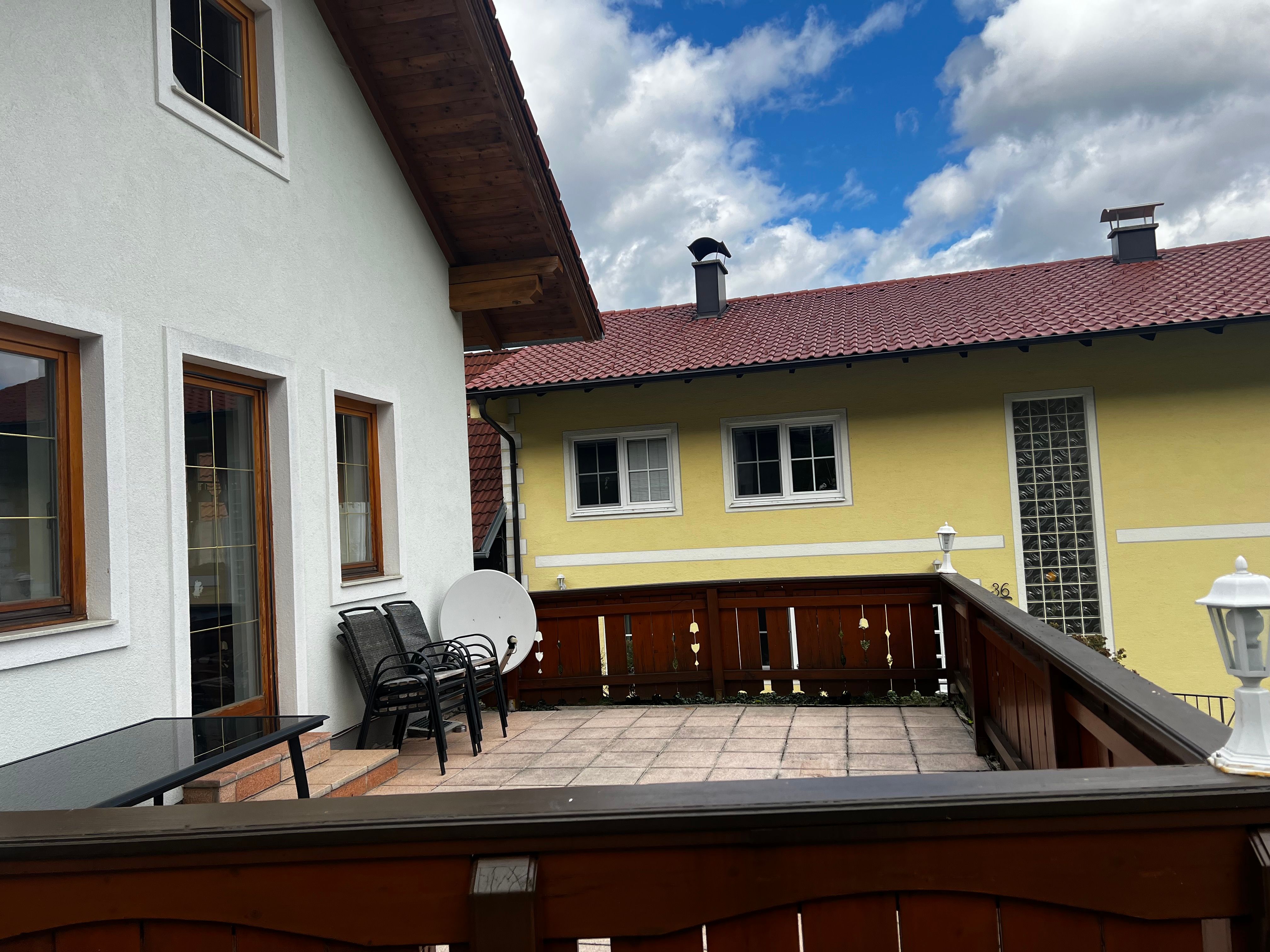 Terasa bytu s dřevěným nábytkem a výhledem na žlutý sousední dům s červenou střechou.