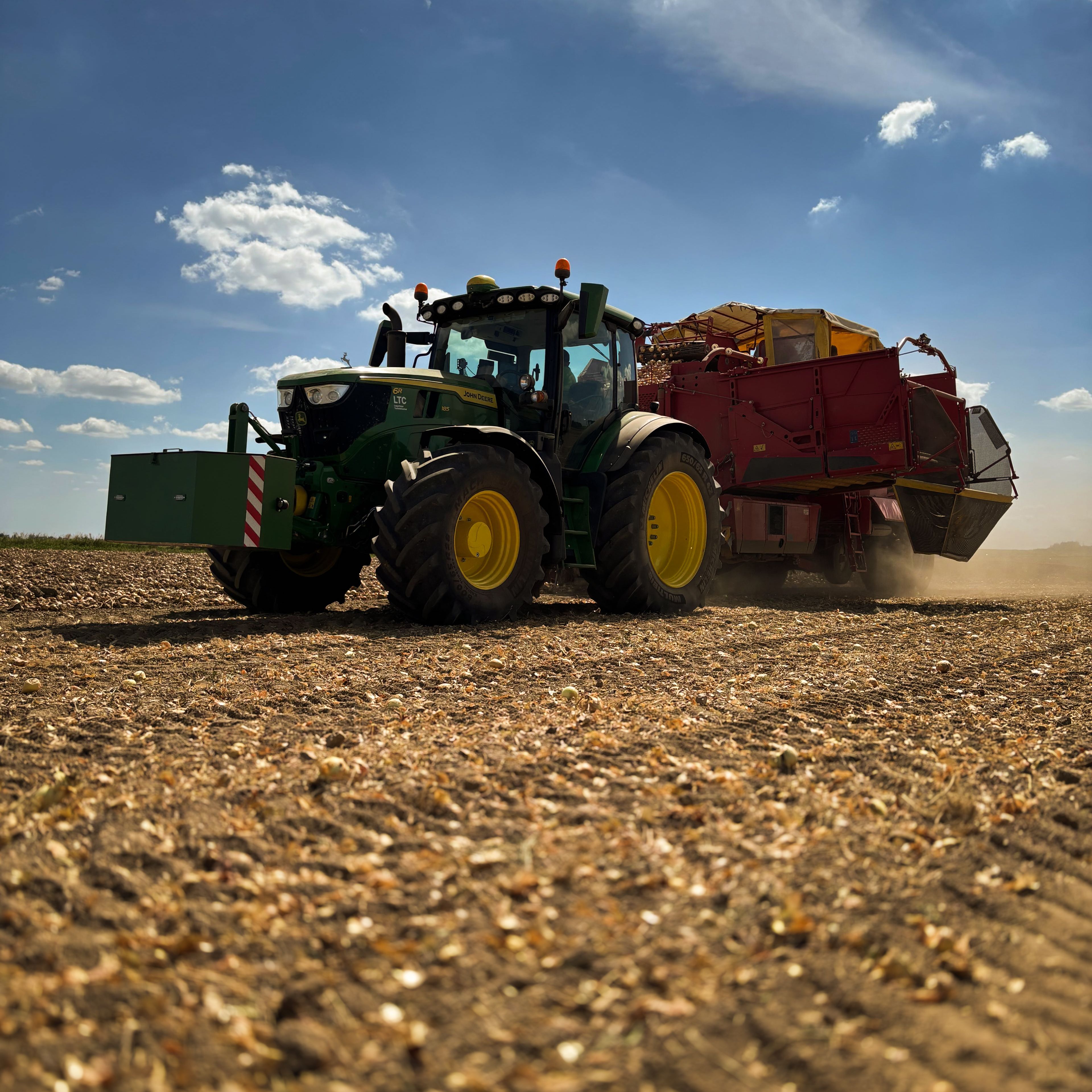 Traktor s přívěsem jede po sklizeném poli pod modrou oblohou s několika mraky.