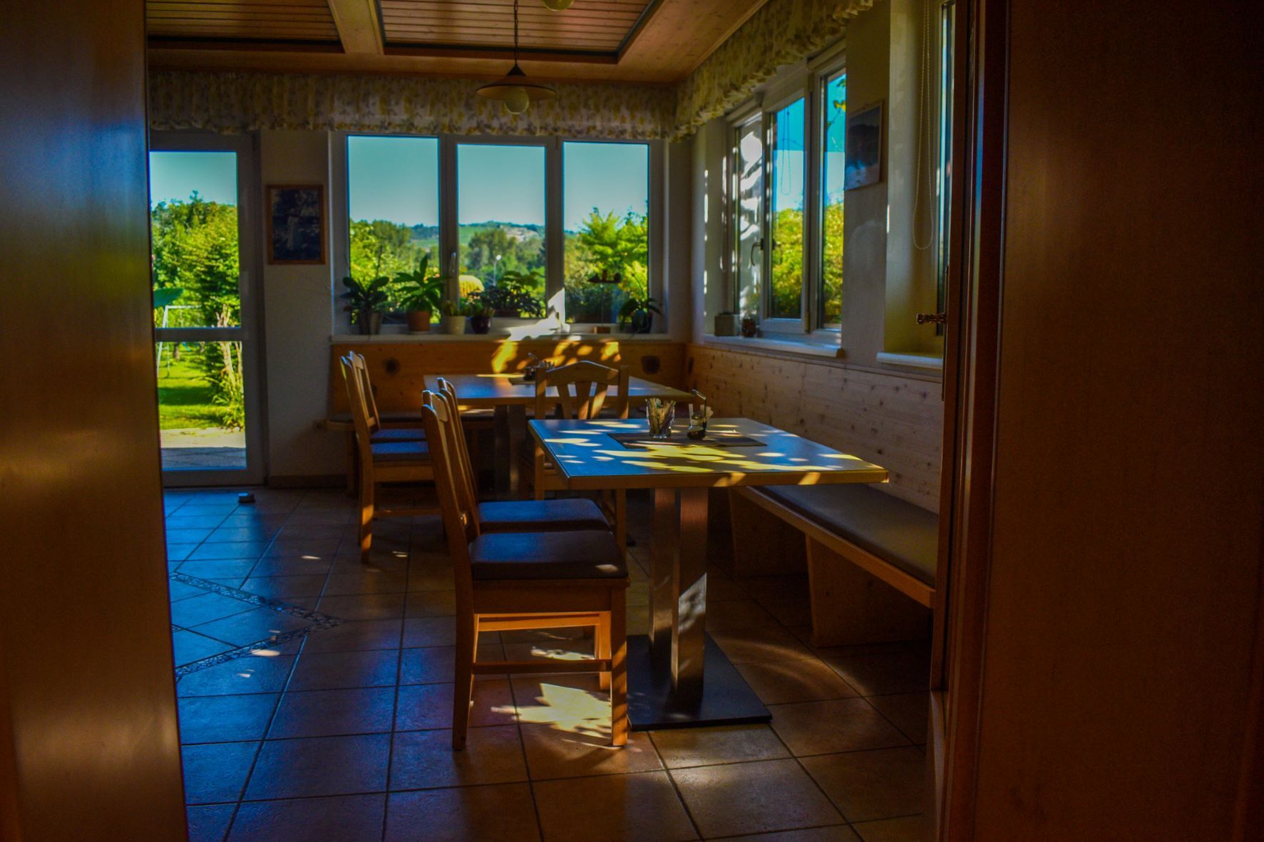 Útulná snídaňová místnost s dřevěným nábytkem a velkými okny propouštějícími sluneční světlo.