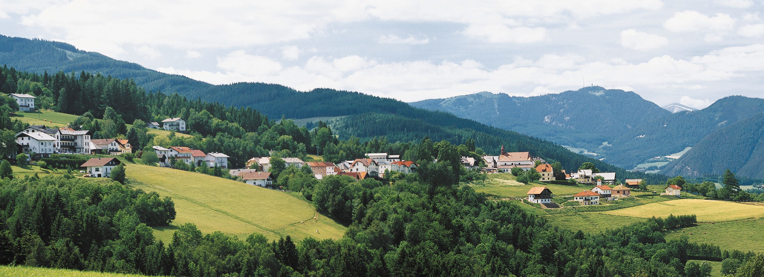 Panorama St. Corona am Wechsel se zelenými kopci a horami v pozadí.