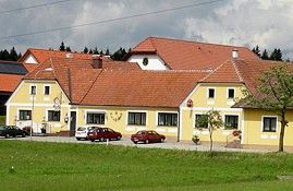 Žlutá budova s červenými střechami a několika auty před ní, obklopená zelenými loukami a stromy.