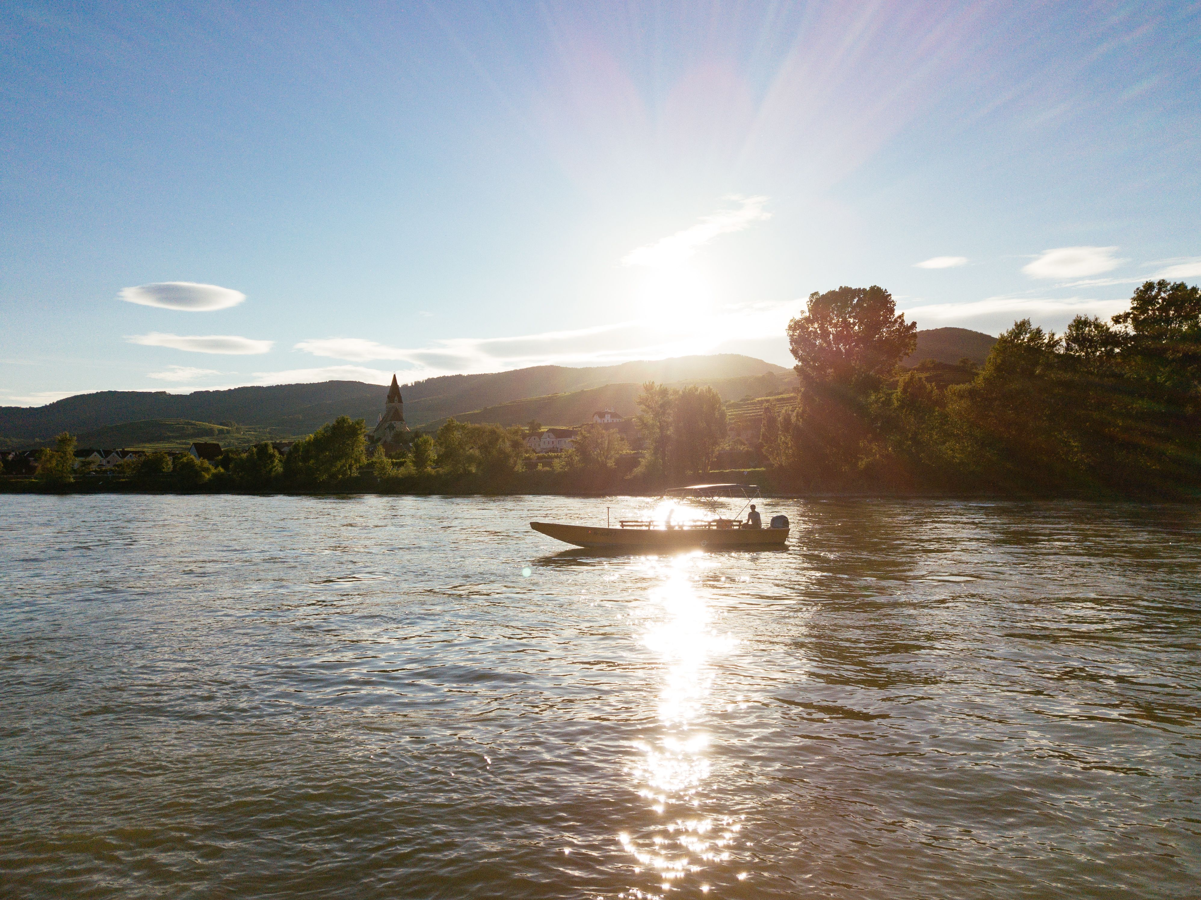 Die sanften Wellen der Donau spiegeln das goldene Licht der untergehenden Sonne wider, während ein Boot gemächlich über das Wasser gleitet. Umgeben von sanften Hügeln und üppigem Grün, bietet diese Szenerie eine perfekte Kulisse für eine entspannende Bootstour. Hier in der Wachau wird der Sommer in seiner schönsten Form erlebbar.