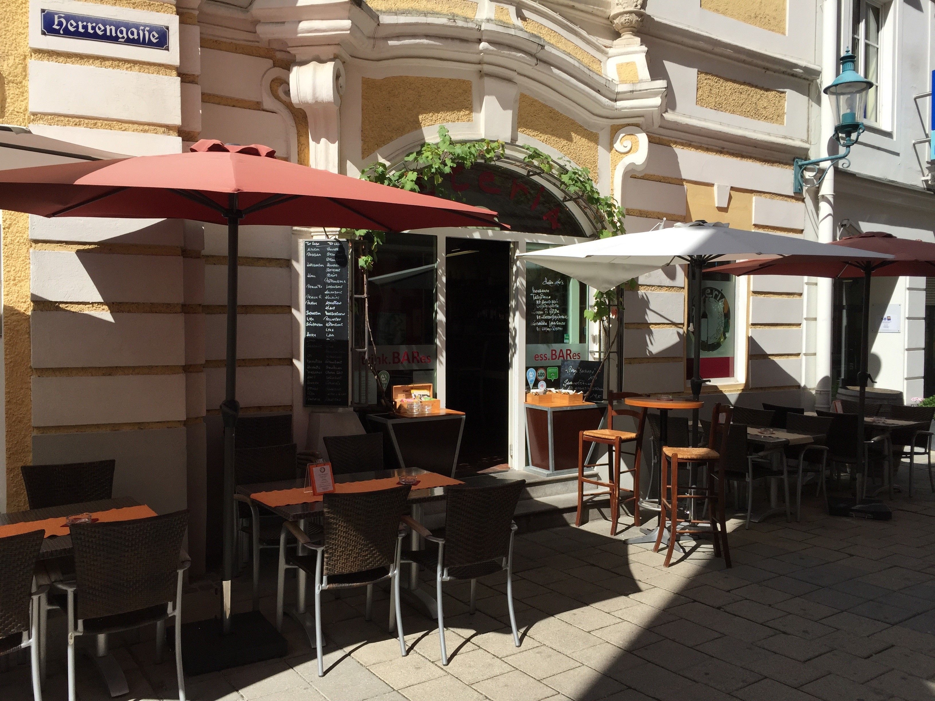 Venkovní prostor restaurace se stoly, židlemi a slunečníky ve slunné uličce.