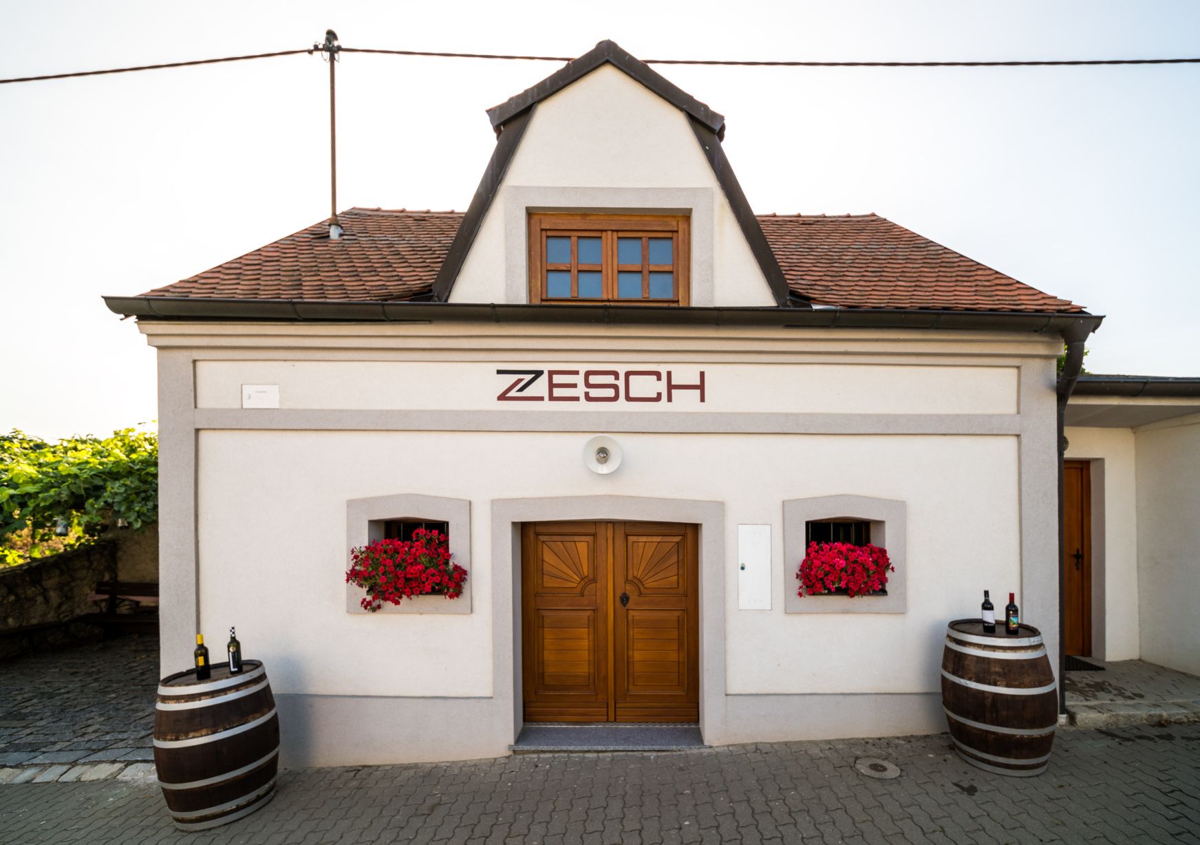 Malá budova s nápisem "Zesch", dva sudy na víno a červené květy.