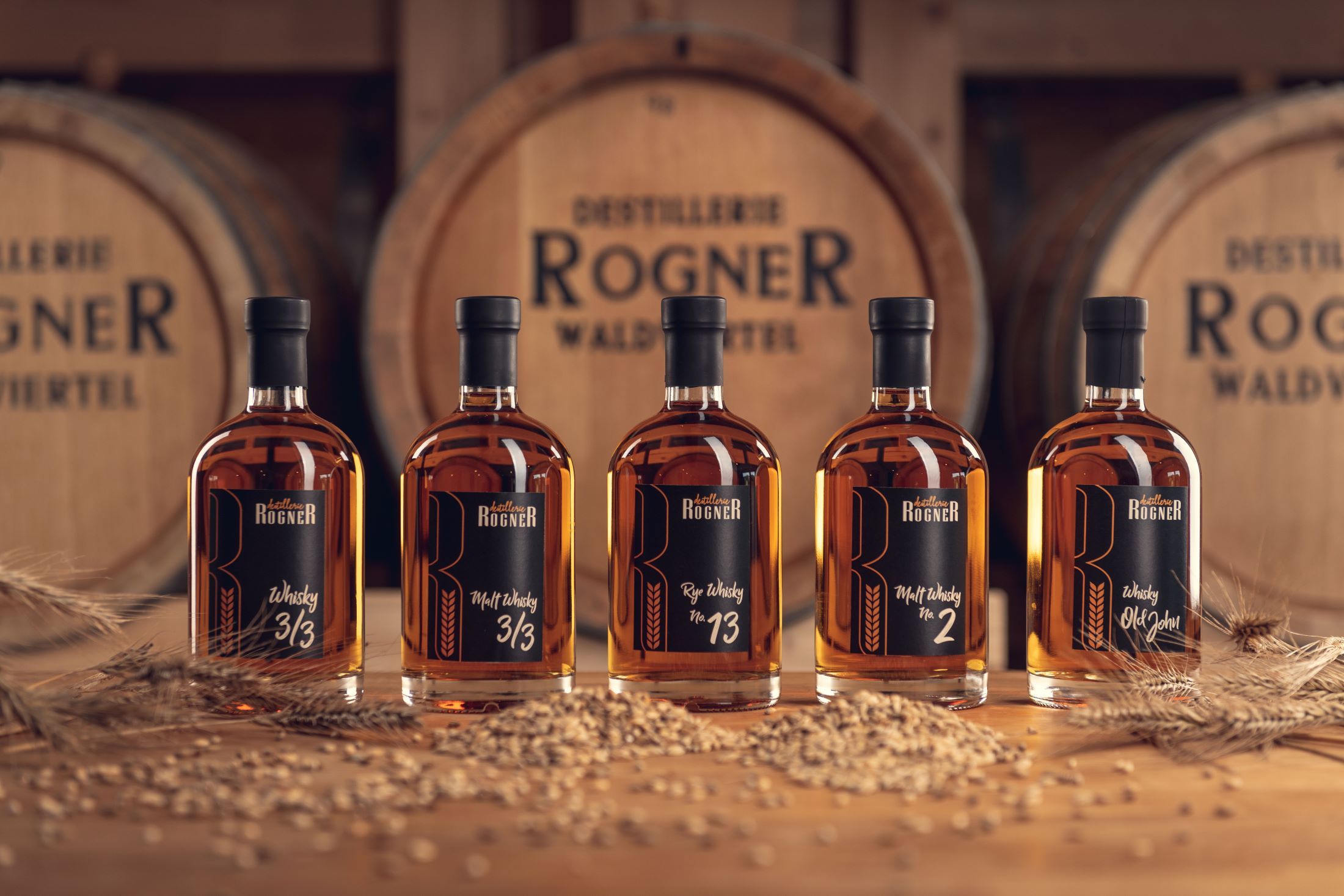 Pět lahví whisky z palírny Rogner před dřevěnými sudy.