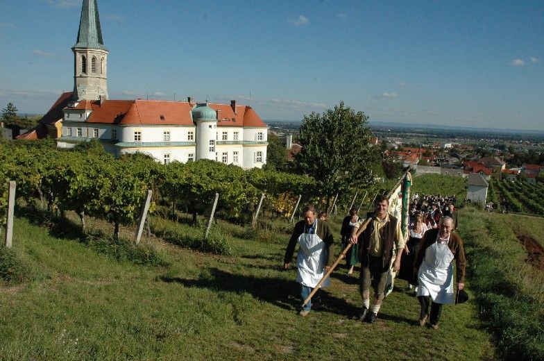 Lidé v tradičních krojích procházejí vinicemi v Gumpoldskirchenu, v pozadí kostel a budovy.
