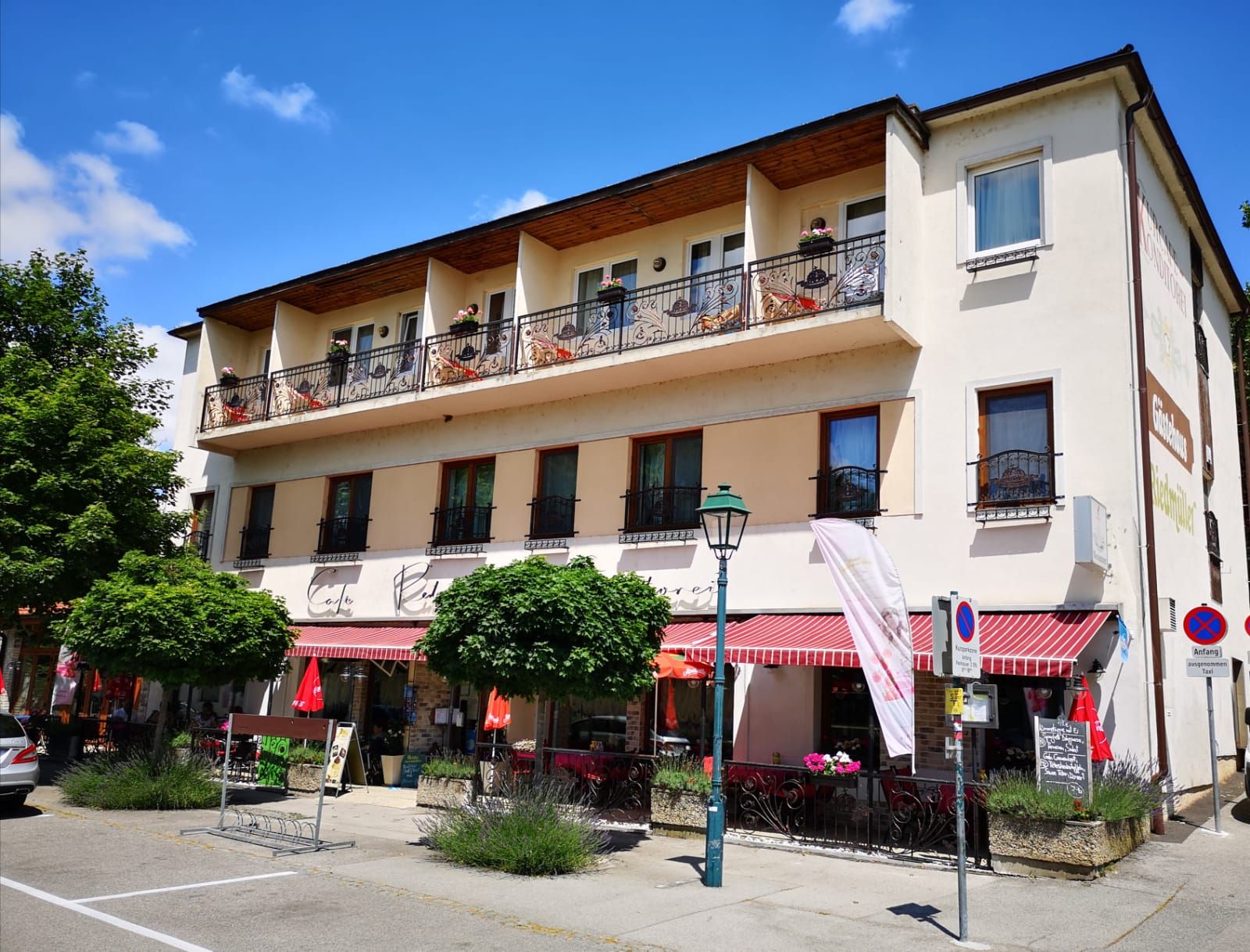 Pohled zvenčí na kavárnu s balkonem a markýzou, obklopenou stromy a dopravními značkami.