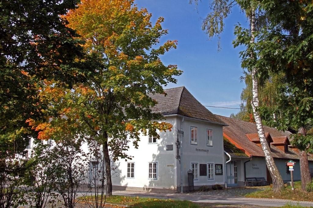 Bílá budova s nápisem "Heimatmuseum", obklopená stromy s podzimním listím.