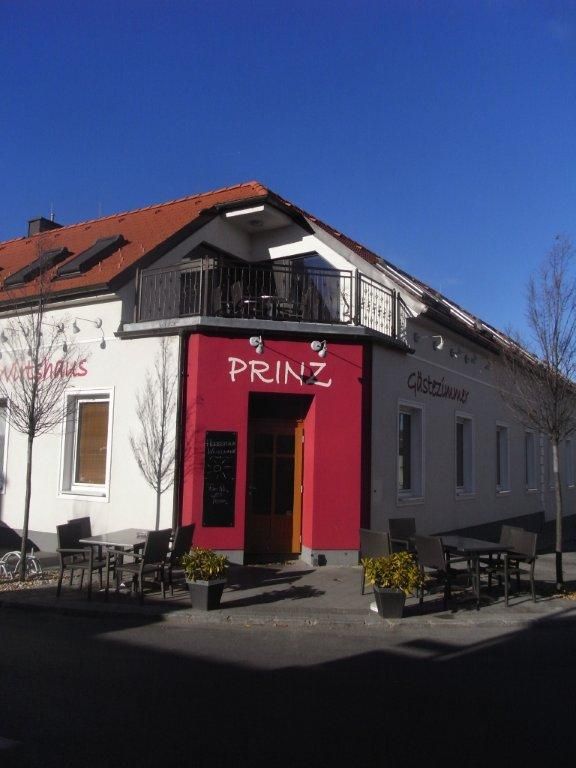 Pohled zvenčí na budovu s označením "Prinz", zahrada pro hosty se stoly a židlemi, modrá obloha.