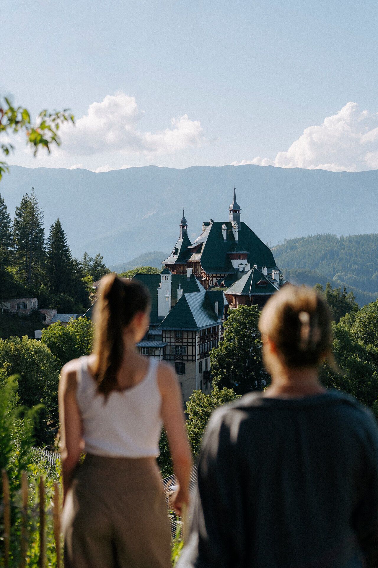 Dva lidé zády ke kameře se dívají na malebnou horskou krajinu. V popředí je vidět hotel Südbahnhotel, zasazený do bujné zeleně. V pozadí se pod jasnou modrou oblohou zvedají hory.