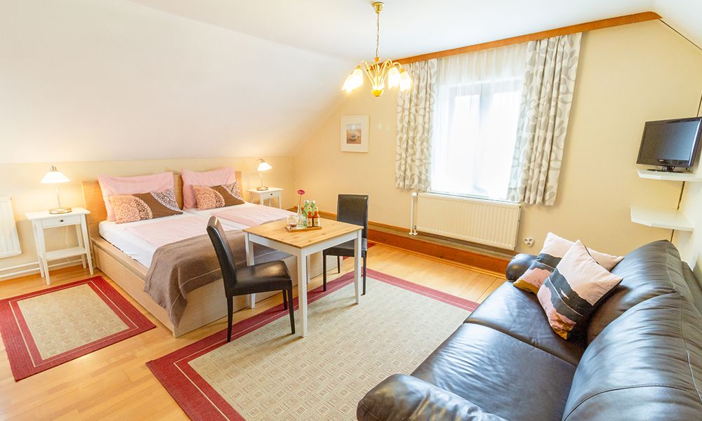 Útulný pokoj pro hosty s manželskou postelí, pohovkou, stolem a židlemi, světlý nábytek.