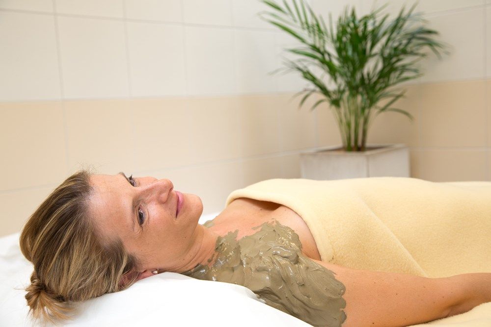 Žena relaxuje se sirným bahnem na rameni, zabalená do deky, ve wellness zóně s rostlinou v pozadí.