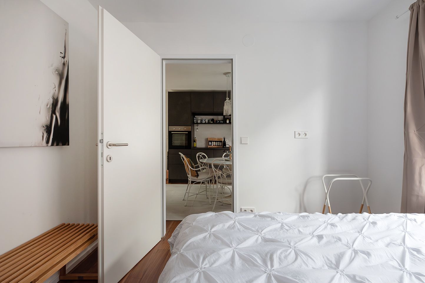 Moderní ložnice s bílým ložním prádlem, otevřenými dveřmi vedoucími do jídelny a minimalistickým nábytkem.