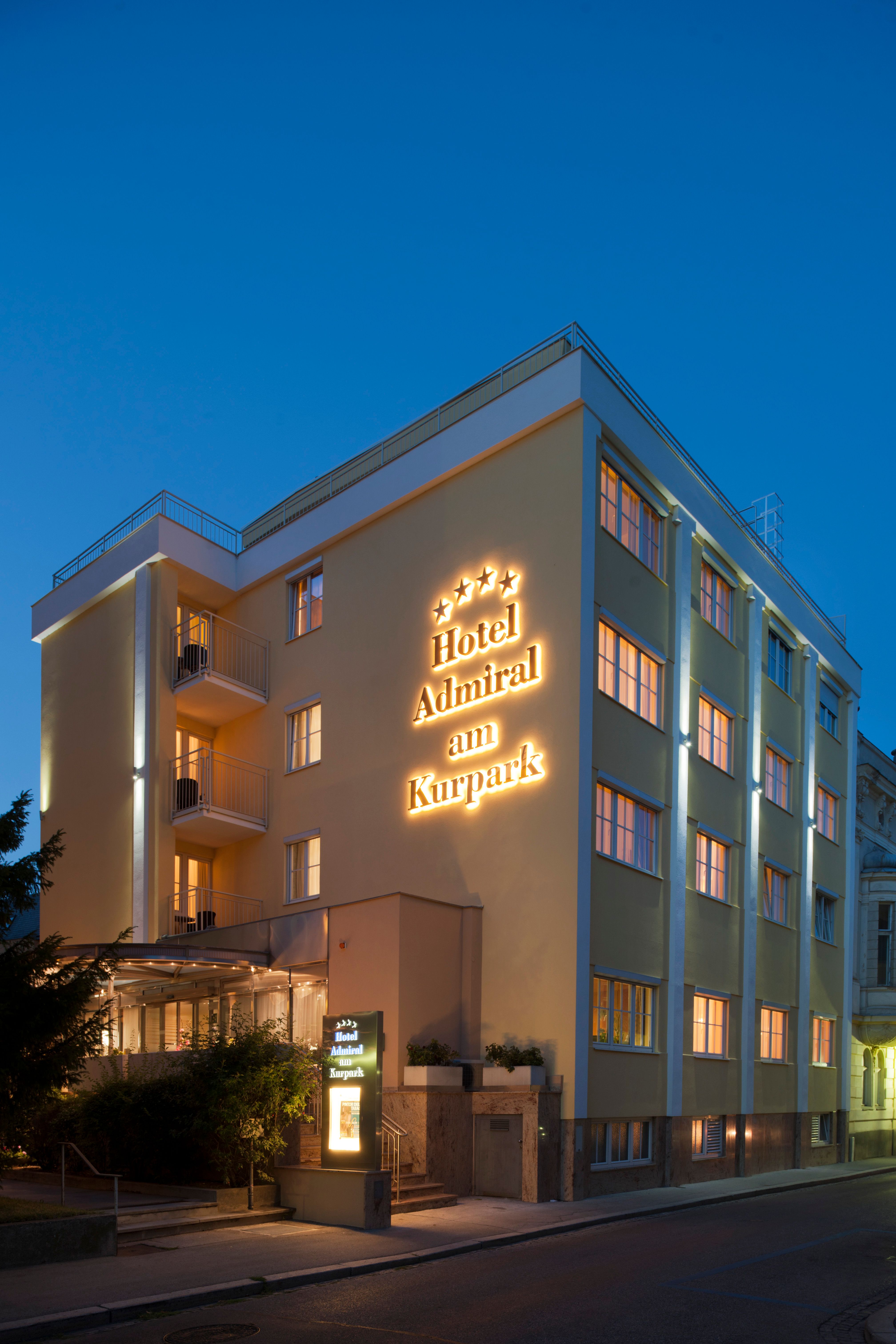 Pohled na exteriér hotelu za soumraku s osvětleným nápisem "Hotel Admiral am Kurpark".