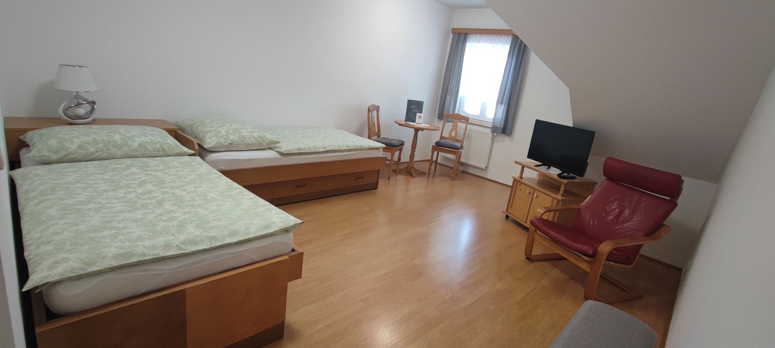 Jednoduše zařízený pokoj se dvěma samostatnými lůžky, malým stolkem a židlemi, televizí a červeným křeslem.