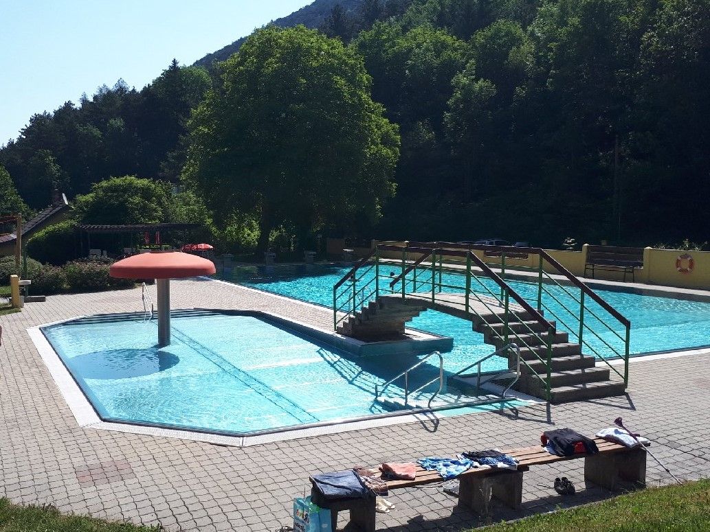 Venkovní bazén s malým bazénem, mostem a hřibovitým vodotryskem. Obklopen stromy a slunečnou oblohou.