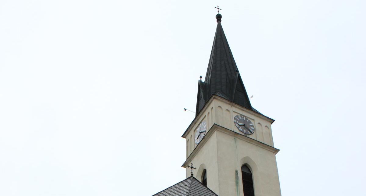 Kostelní věž s hodinami a špičatou střechou před jasnou oblohou.