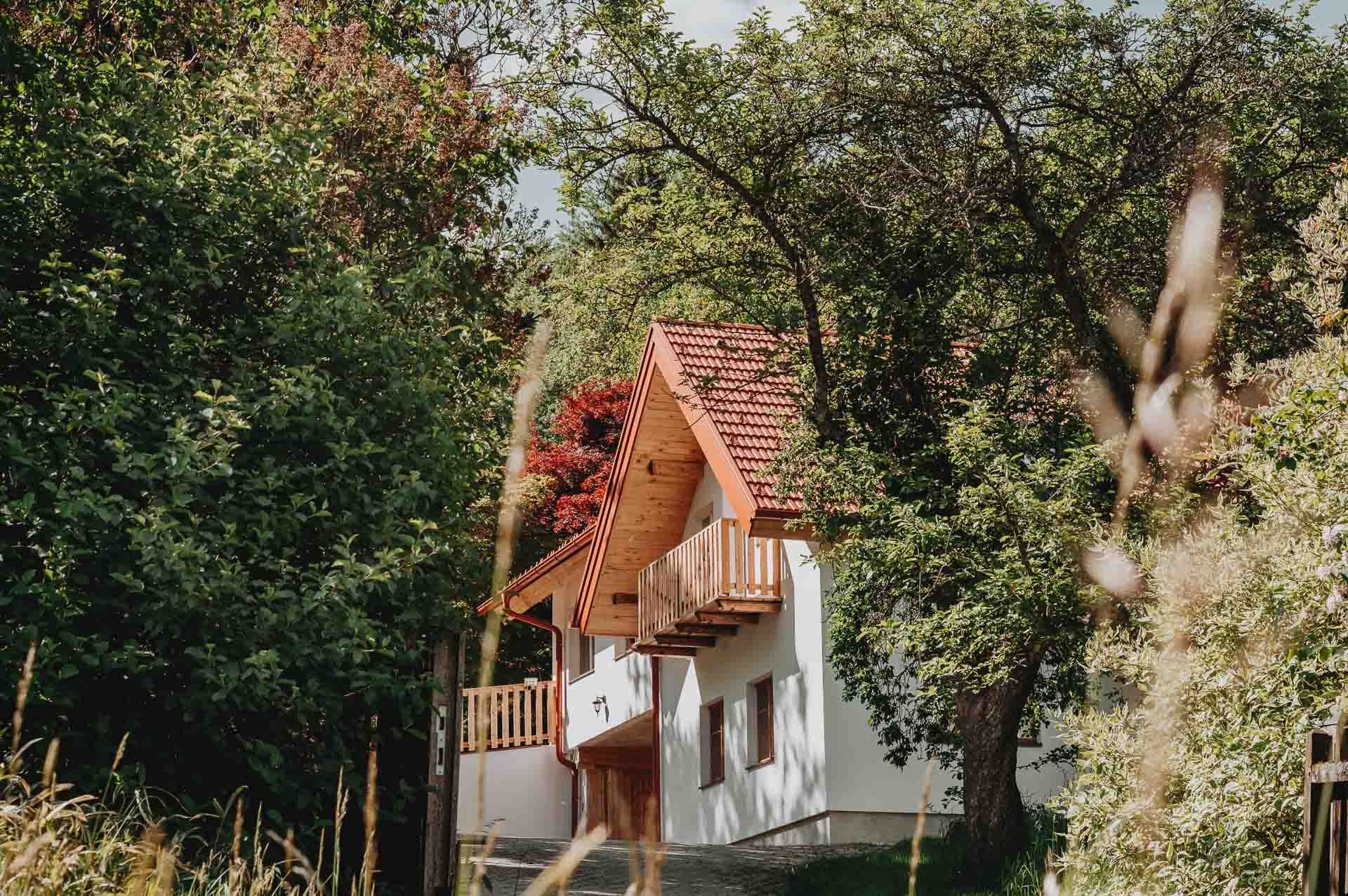 Bílý dům s červenou taškovou střechou, obklopený stromy a rostlinami.