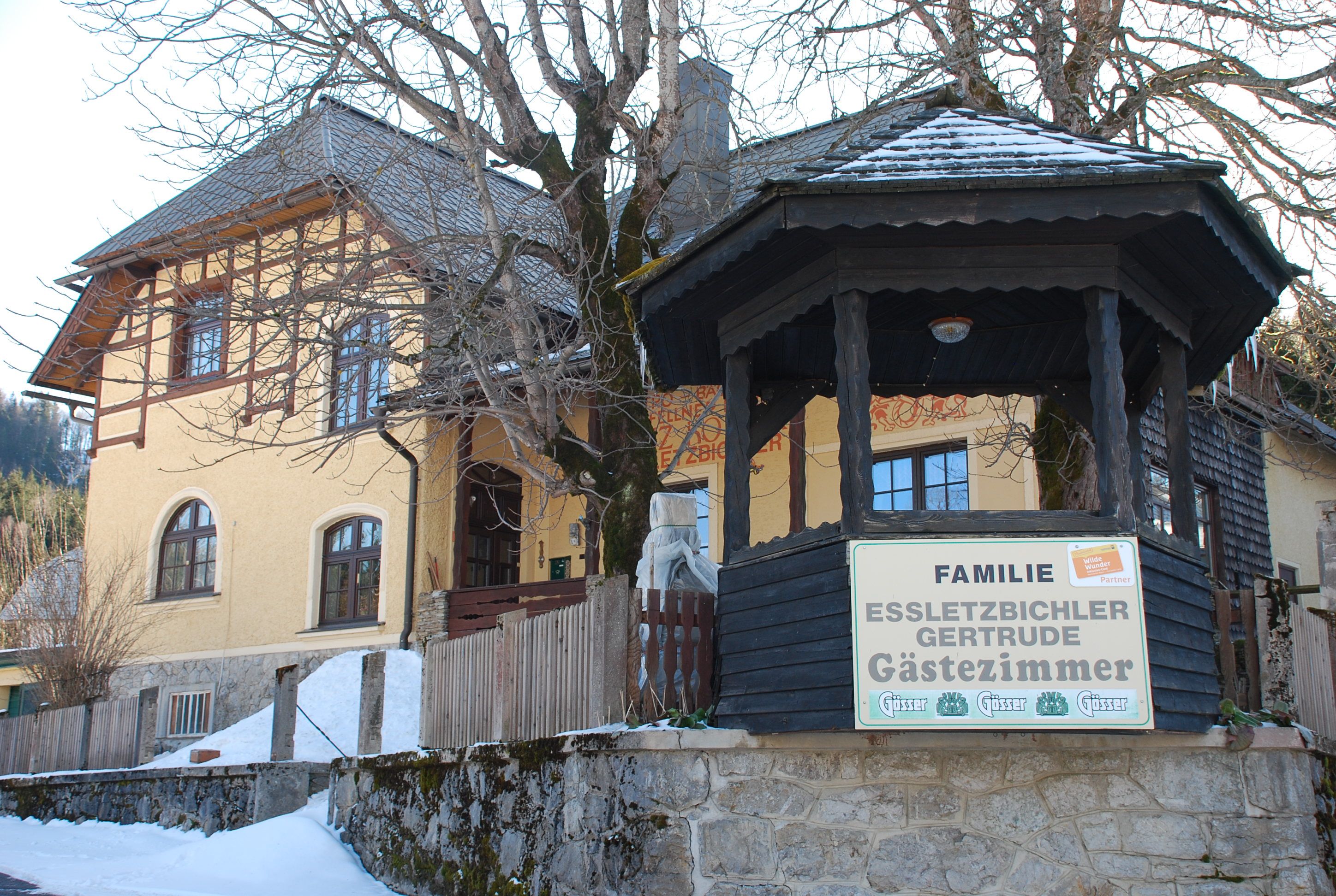 Žlutý dům pro hosty s dřevěnou verandou a nápisem "Familie Essletzbichler Gertrude Gästezimmer".
