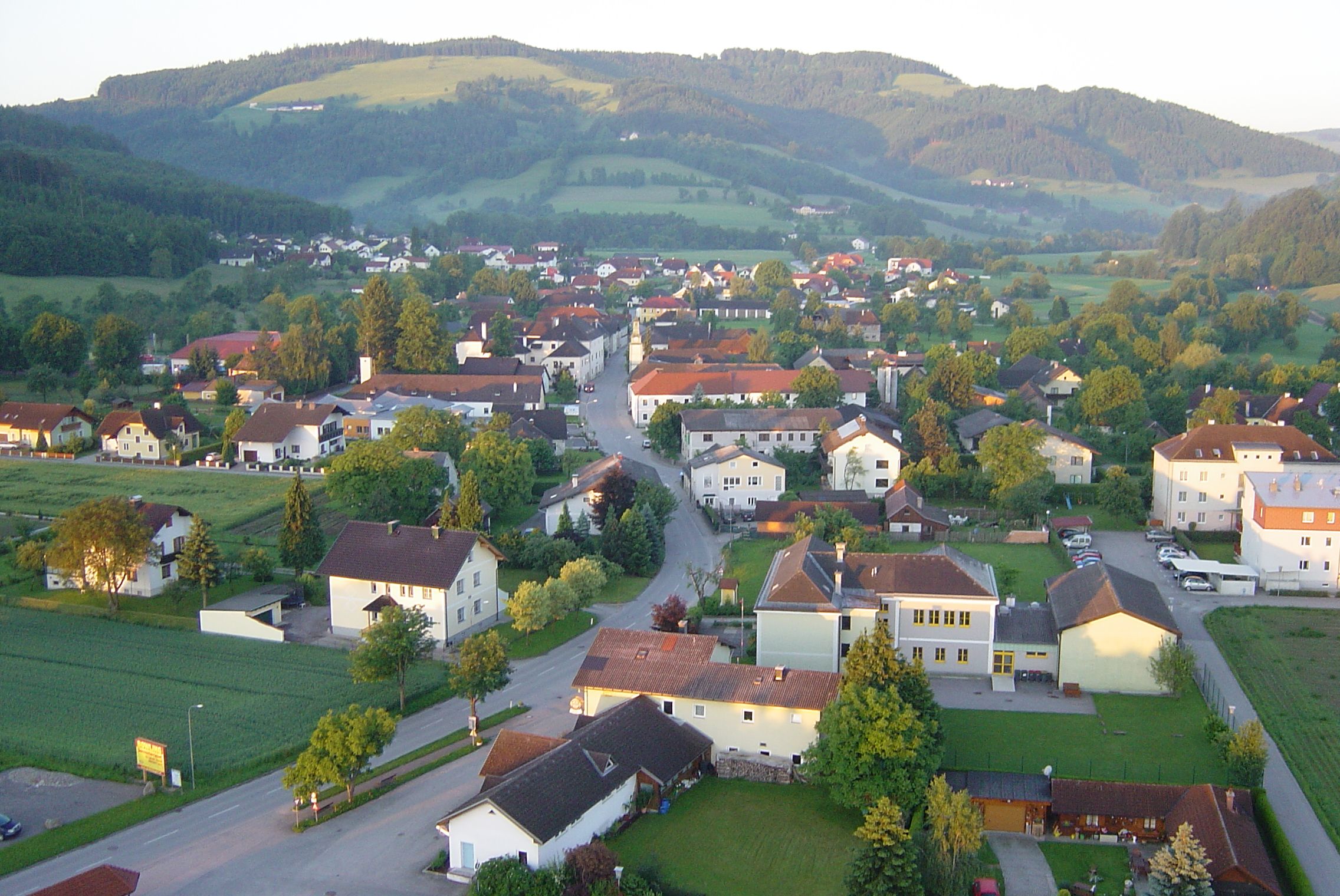 Letecký pohled na vesnici s domy, ulicemi a okolními kopci v pozadí.