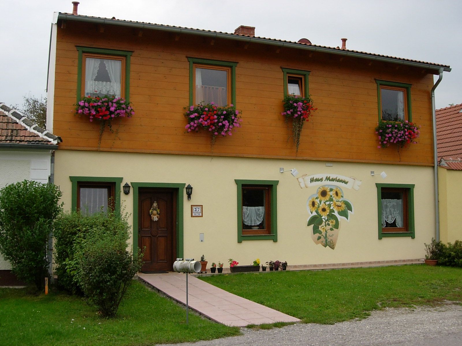 Dvoupatrový dům s dřevěným obložením a květinovými truhlíky, označený jako "Haus Marianne".