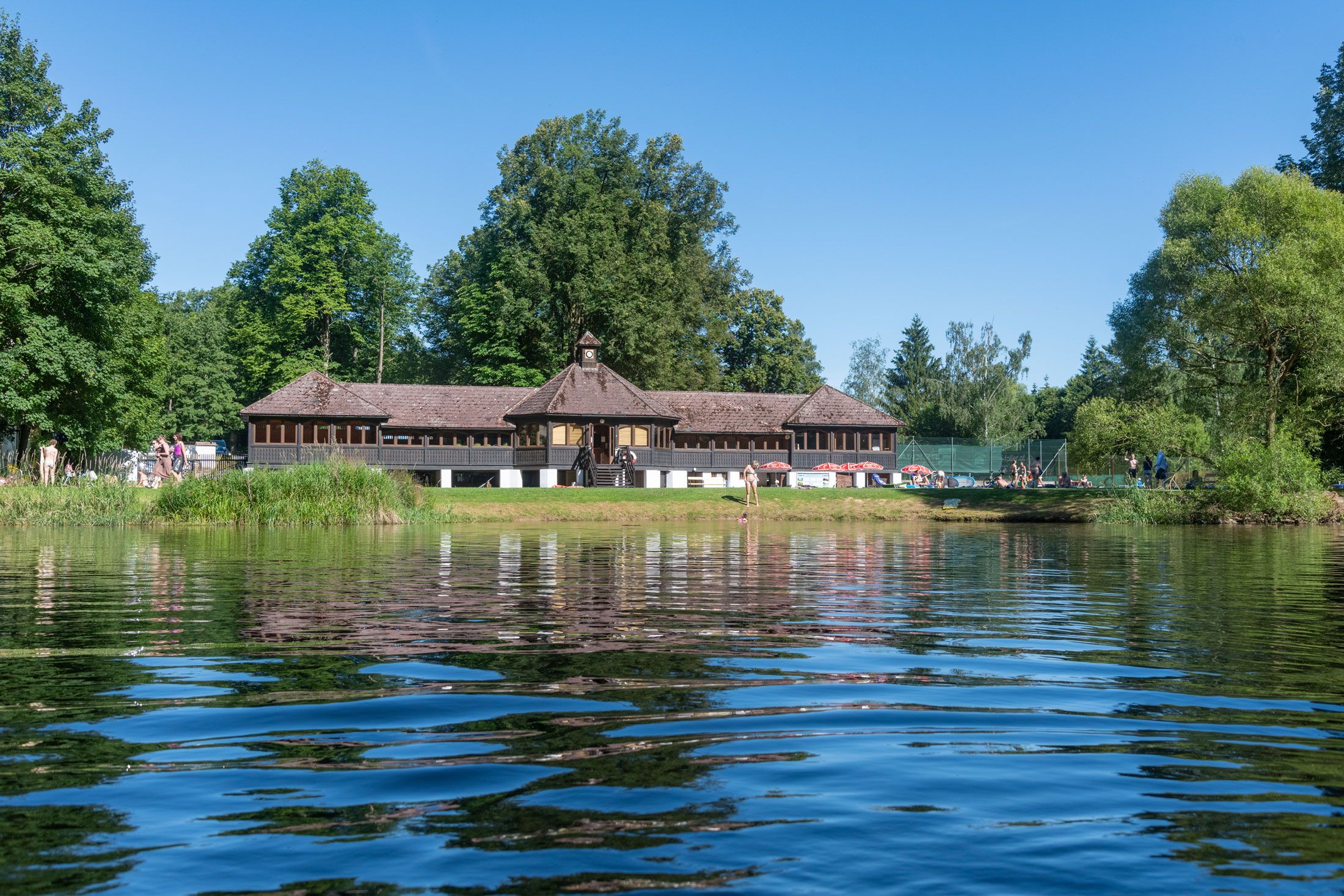 Budova na břehu jezera se stromy v pozadí a lidmi užívajícími si slunečného počasí.