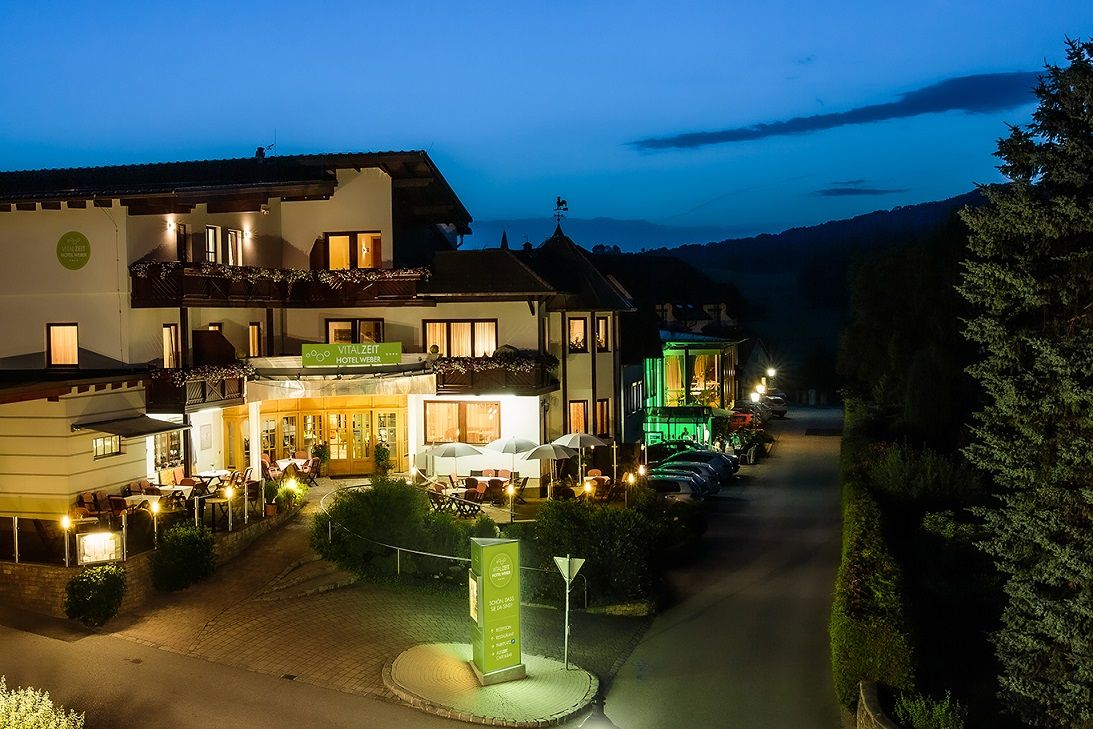 Pohled na noční, teplým světlem osvětlený exteriér hotelu Vitalzeit Weber, obklopený stromy a zaparkovanými auty.