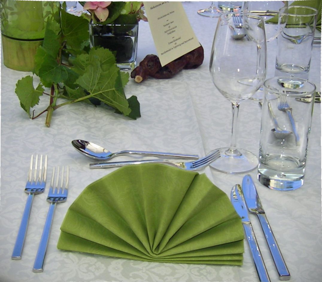 Elegantní dekorace stolu se zeleným skládaným ubrouskem, příbory, sklenicí na víno a sklenicí na vodu na bílém ubrusu.