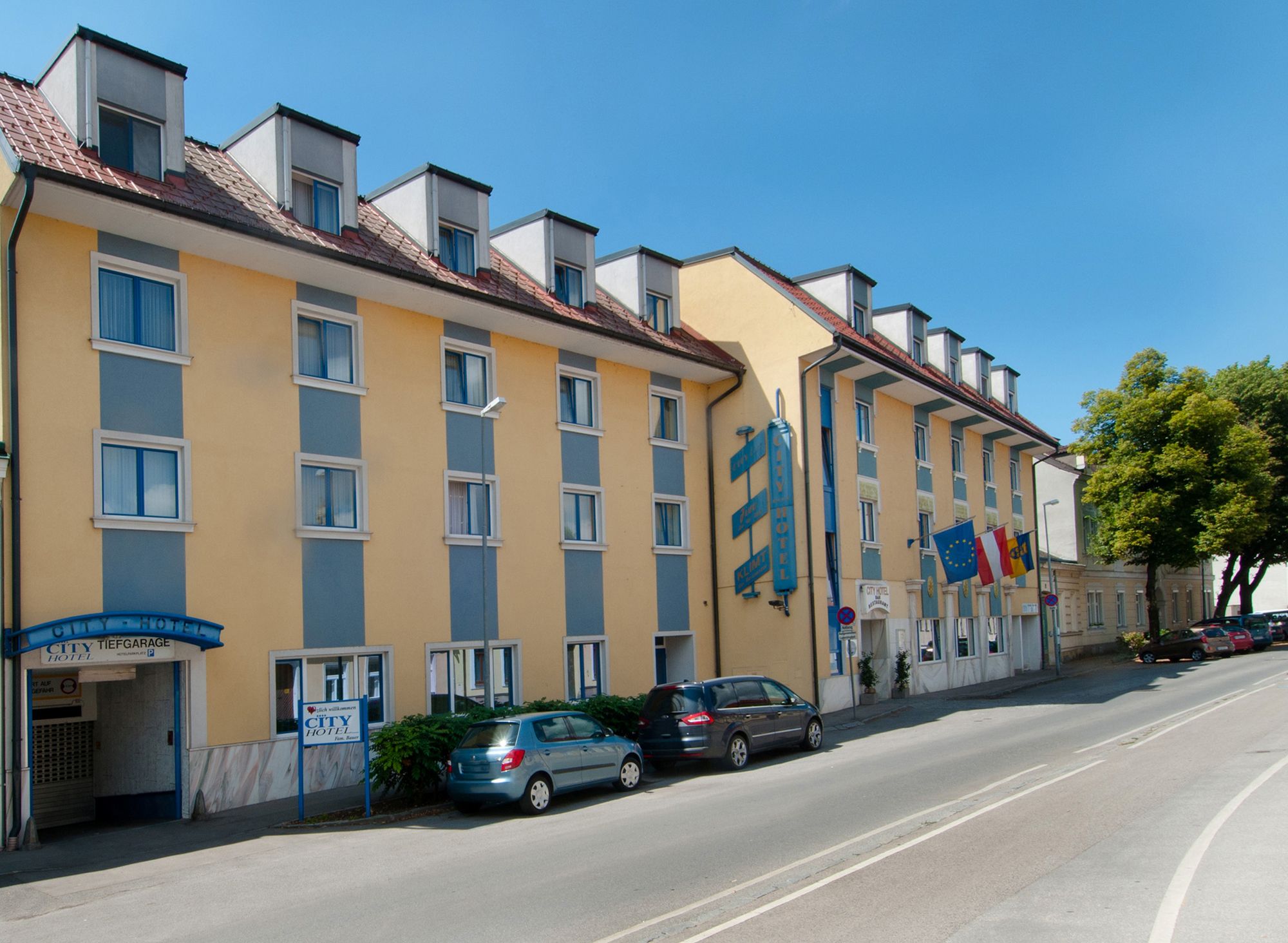 Pohled zvenčí na žlutomodrý hotel s několika okny a zaparkovanými auty před ním.