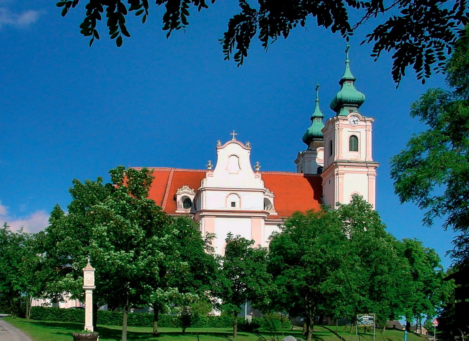 Maria Dreieichen bazilika s červenými střechami a zelenými věžemi, obklopená stromy.
