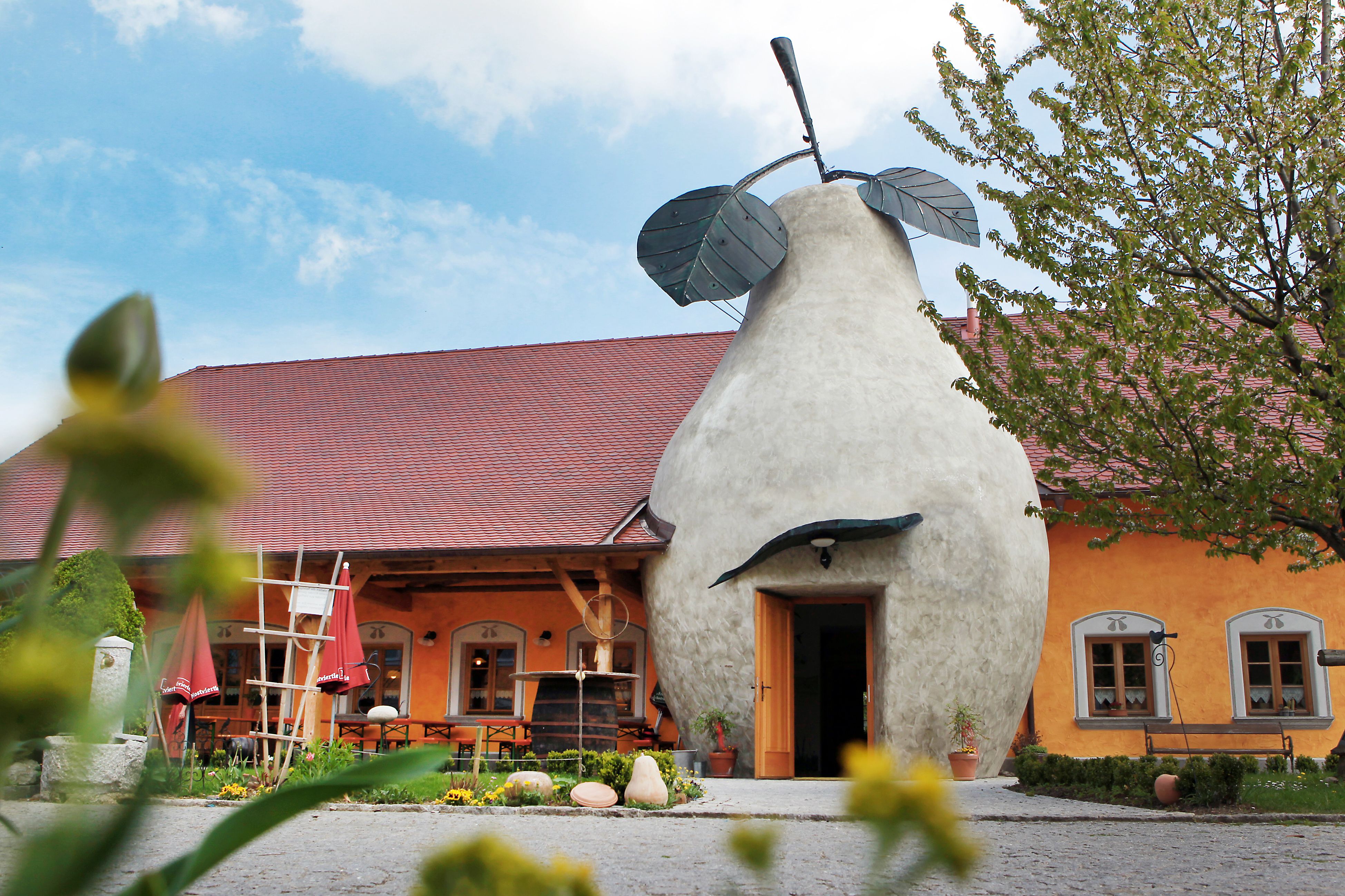 Budova s vchodem ve tvaru hrušky a červenou střechou, obklopená rostlinami.